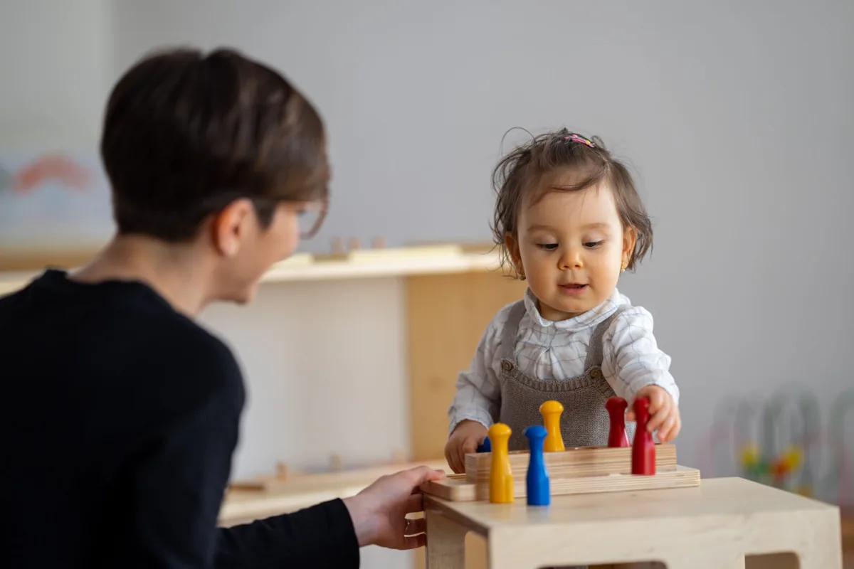 Una niña manipula piezas de colores sobre una base de madera junto a una adulta.