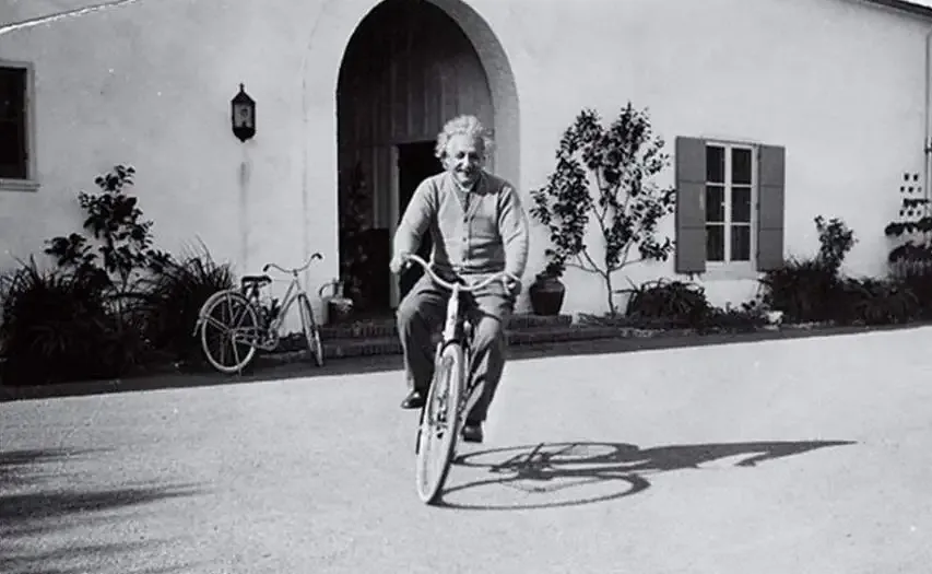 Albert Einstein andando en bicicleta frente a una casa.